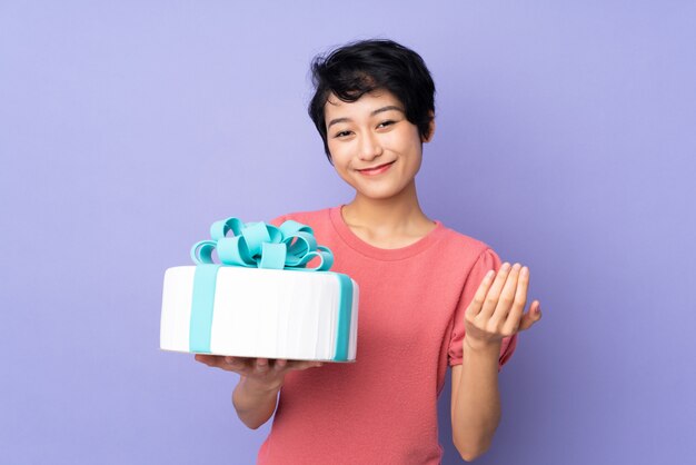 Young Vietnamese woman with short hair holding a big cake over purple wall inviting to come with hand