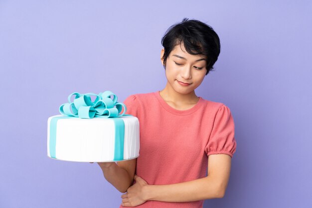 Young Vietnamese woman with short hair holding a big cake over purple wall with sad expression