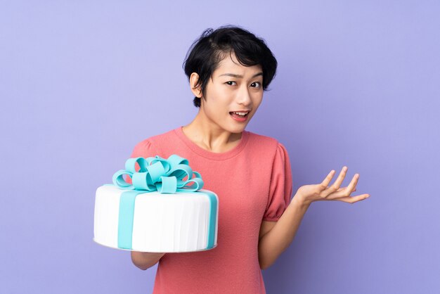 Young Vietnamese woman with short hair holding a big cake over purple wall with shocked facial expression