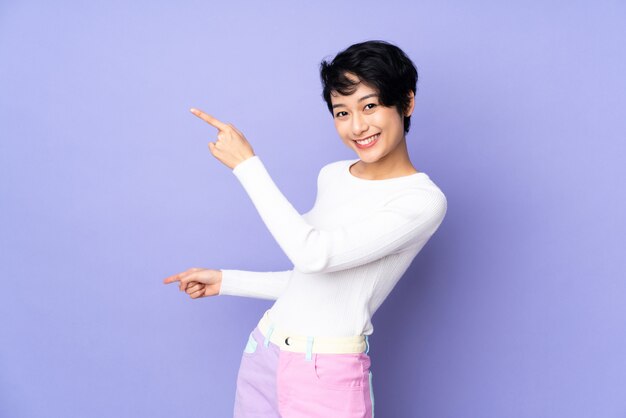 Young Vietnamese woman with short hair over isolated purple wall pointing finger to the side and presenting a product