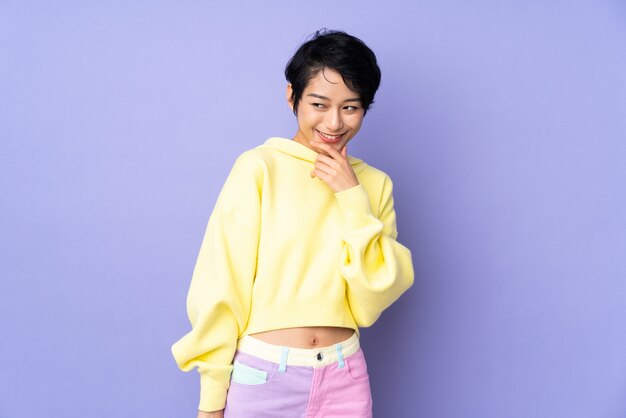 Young Vietnamese woman with short hair over purple wall looking to the side and smiling