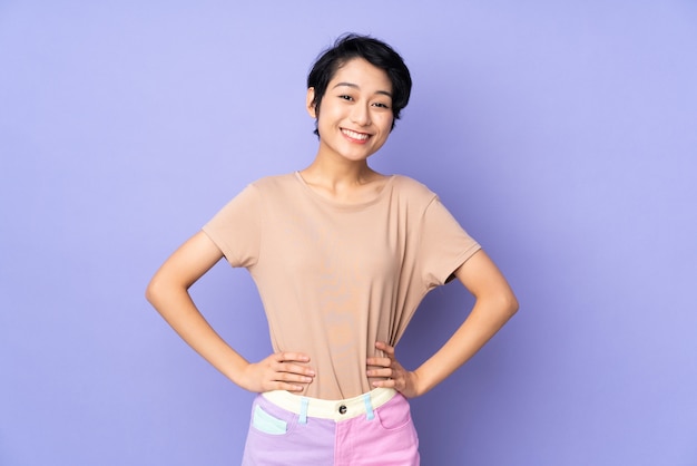 Young Vietnamese woman with short hair over purple wall posing with arms at hip and smiling