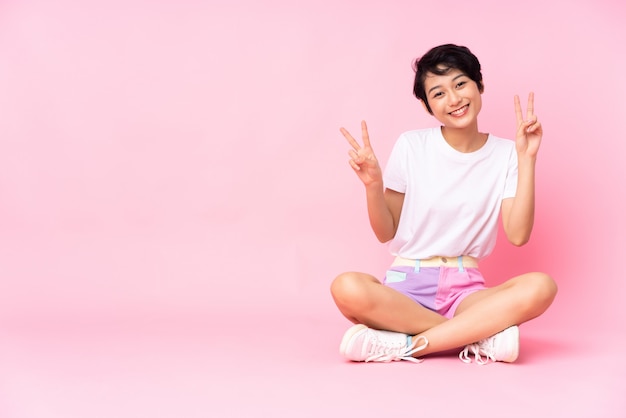 Young Vietnamese woman with short hair sitting on the floor over isolated pink wall showing victory sign with both hands