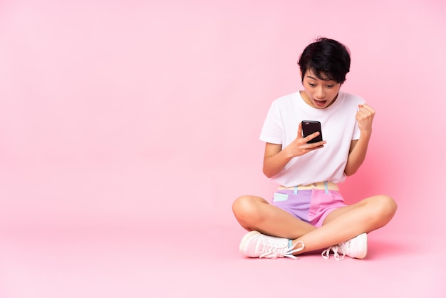 Young Vietnamese woman with short hair sitting on the floor over isolated pink wall surprised and sending a message