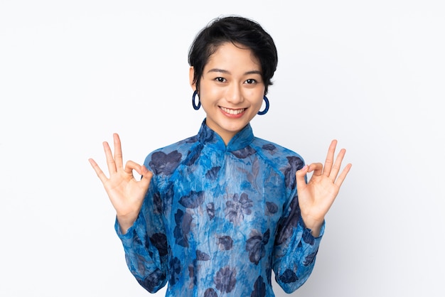 Young Vietnamese woman with short hair wearing a traditional dress over white wall showing ok sign with two hands