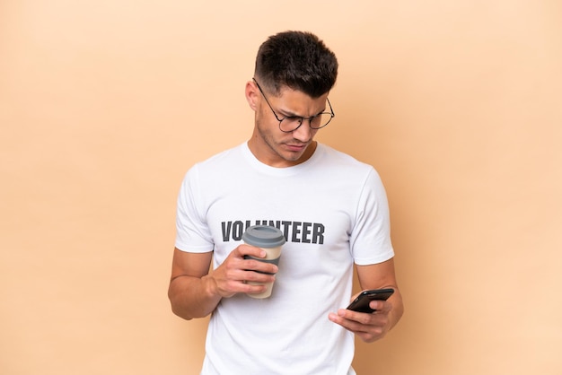 Young volunteer caucasian man isolated on beige background holding coffee to take away and a mobile