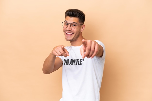 Young volunteer caucasian man isolated on beige background points finger at you while smiling