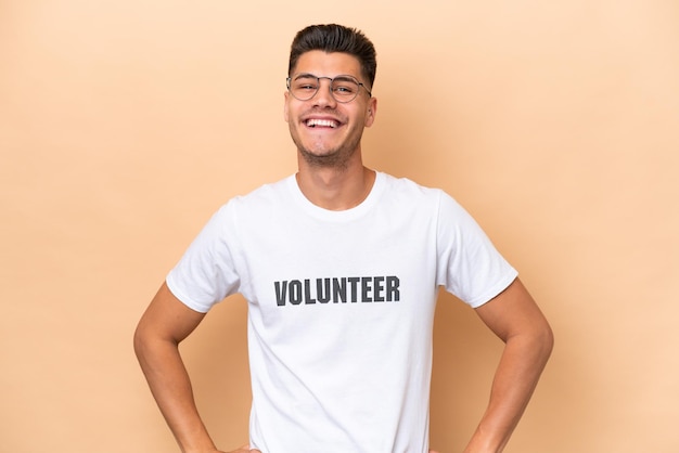 Young volunteer caucasian man isolated on beige background posing with arms at hip and smiling