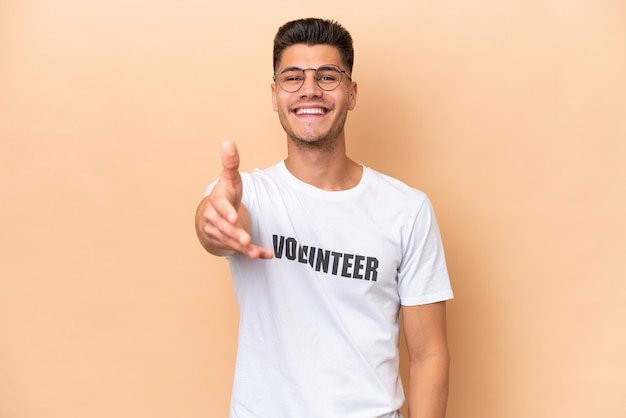 Young volunteer caucasian man isolated on beige background shaking hands for closing a good deal