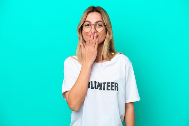 Young volunteer Uruguayan woman isolated on blue background happy and smiling covering mouth with hand