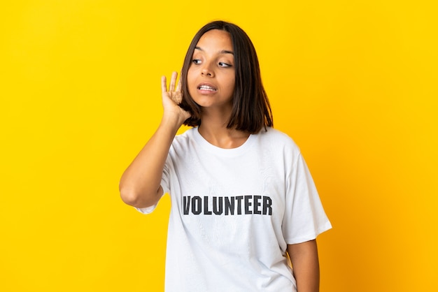 Young volunteer woman isolated on yellow wall listening to something by putting hand on the ear