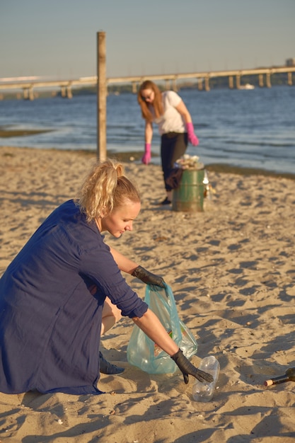 Young volunteers in black gloves are walking with garbage bags along a dirty beach of the river