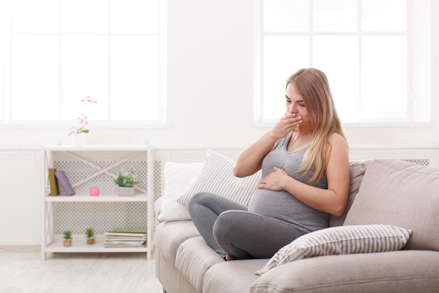 Young vomiting woman sitting on sofa. Pregnant blonde suffering with nausea. Pregnancy symptoms, expectation, parenthood concept