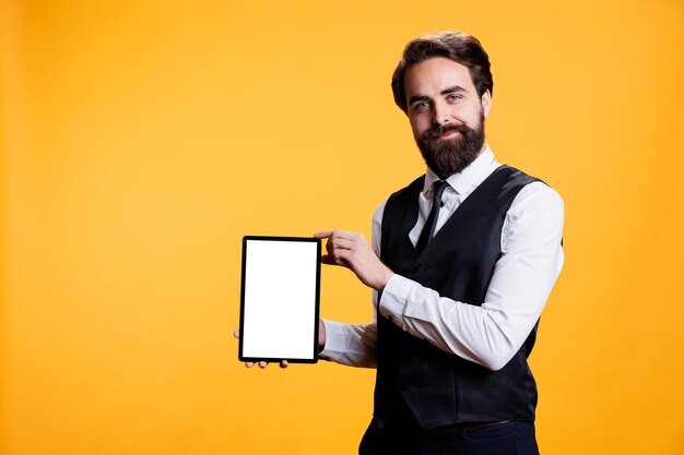 Young waiter holding tablet with white screen on camera, presenting isolated empty template against yellow background. professional employee shows device with blank mockup display