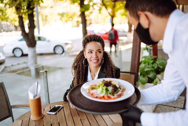 Young waiter in protective face mask and gloves with ordered meals ready to serving guest Covid2019