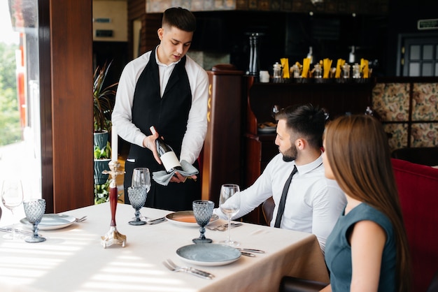 A young waiter in a stylish apron demonstrates and offers a fine wine to a beautiful couple in a restaurant. Customer service.