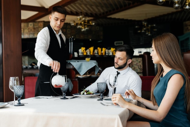 A young waiter in a stylish apron serves a table with a\
beautiful couple in an elegant restaurant customer service in an\
elite restaurant and public catering establishment