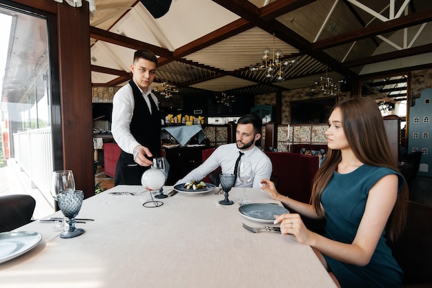 A young waiter in a stylish apron serves a table with a
beautiful couple in an elegant restaurant customer service in an
elite restaurant and public catering establishment