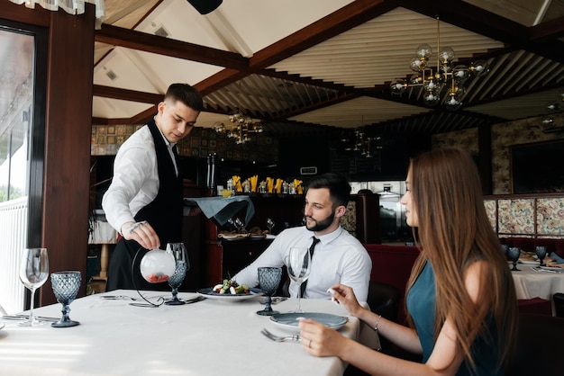 スタイリッシュなエプロンを着た若いウェイターが、エレガントなレストランで美しいカップルと一緒にテーブルを提供します。エリートレストランと公共のケータリング施設でのカスタマーサービス
