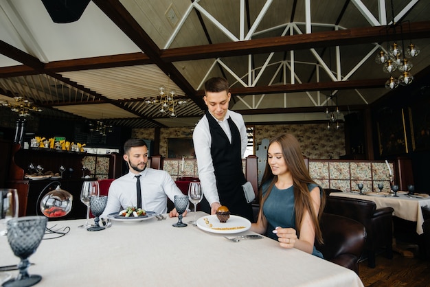 A young waiter in a stylish apron serves a table with a beautiful couple in a refined restaurant. Customer service.