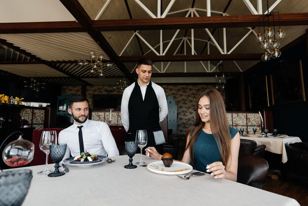 A young waiter in a stylish apron serves a table with a\
beautiful couple in a refined restaurant exquisite delicacies of\
haute cuisine closeup