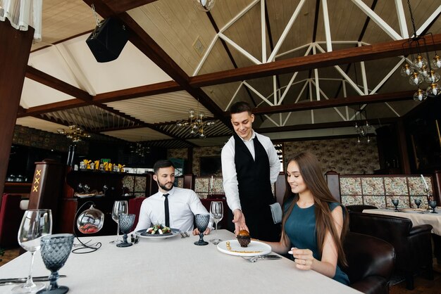 A young waiter in a stylish apron serves a table with a\
beautiful couple in a refined restaurant exquisite delicacies of\
haute cuisine closeup