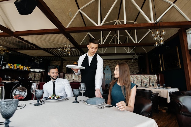 A young waiter in a stylish apron serves a table with a beautiful couple in a refined restaurant Exquisite delicacies of haute cuisine closeup