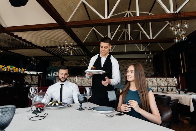 A young waiter in a stylish apron serves a table with a beautiful couple in a refined restaurant Exquisite delicacies of haute cuisine closeup