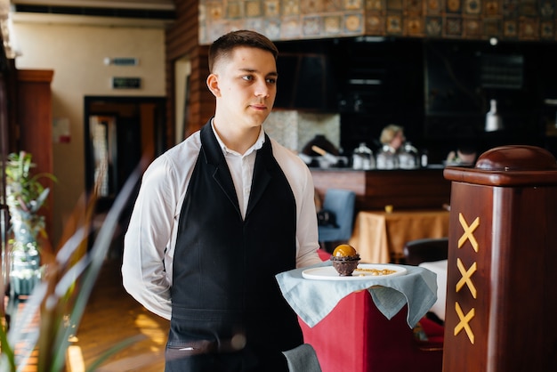 Молодой официант в стильной униформе стоит с изысканным блюдом на подносе возле стола в красивом ресторане крупным планом. Ресторанная деятельность самого высокого уровня.