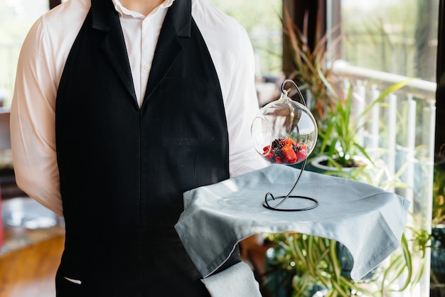 A young waiter in a stylish uniform stands with an exquisite\
dish on a tray near the table in a beautiful restaurant close-up.\
restaurant activity, of the highest level.