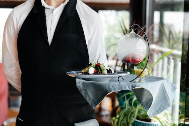 A young waiter in a stylish uniform stands with an exquisite dish on a tray near the table in a beautiful restaurant closeup Restaurant activity of the highest level