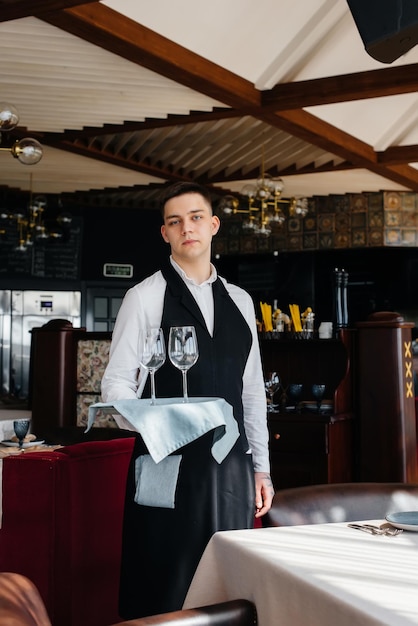 Молодой официант в стильной униформе стоит с бокалами на подносе возле стола в красивом ресторане для гурманов крупным планом. Ресторанная деятельность самого высокого уровня.