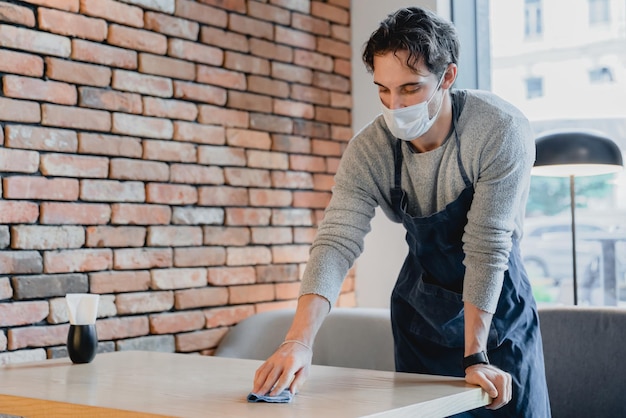 Young waiter wearing medical mask against coronavirus wiping\
table with cloth in loft coffee shop