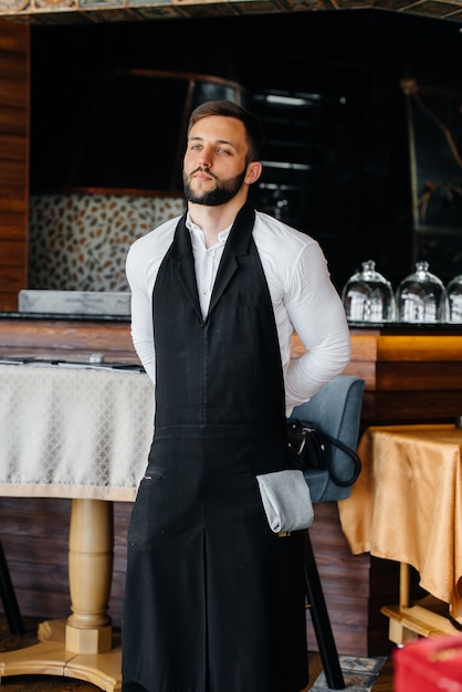 A young waiter with a beard puts on an apron and prepares for a working day in a fine restaurant.