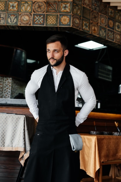 A young waiter with a beard puts on an apron and prepares for a working day in a fine restaurant