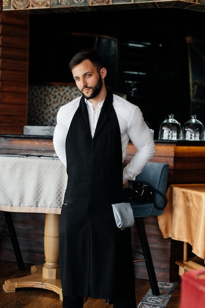 A young waiter with a beard puts on an apron and prepares for a working day in a fine restaurant