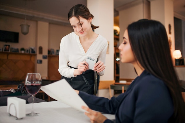 Young waitress stand besides sitting customer and look at menu she point