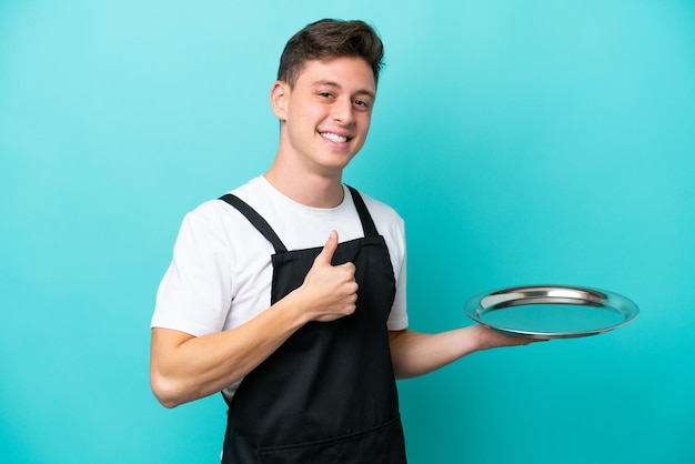 Young waitress with tray isolated on blue background giving a thumbs up gesture