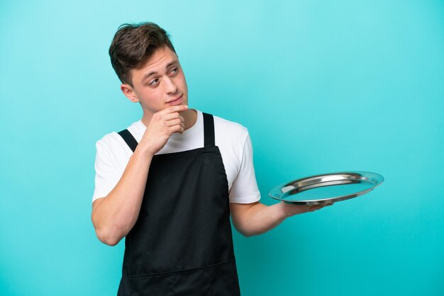 Young waitress with tray isolated on blue background and looking up