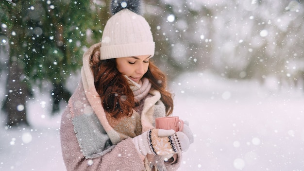 暖かい服を着た若い女性は、雪の降る冬の日に手に温かい飲み物を持っています