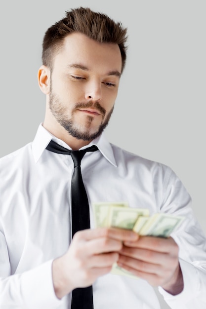 Young and wealthy. Confident young man in shirt and tie counting money and while standing against grey background