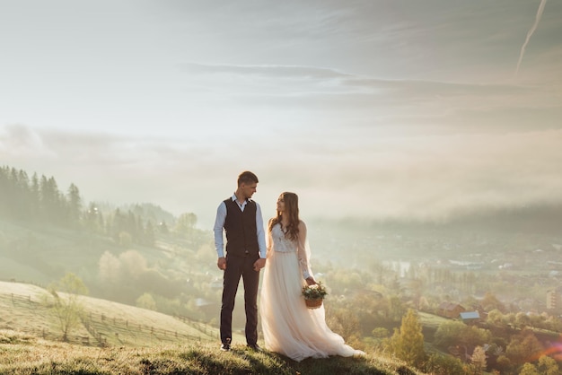 Young wedding couple waking on the hill in the mountains with beautiful landscape and fog on the background at sunrise. Back view stylish couple of bride and groom in a wedding walk