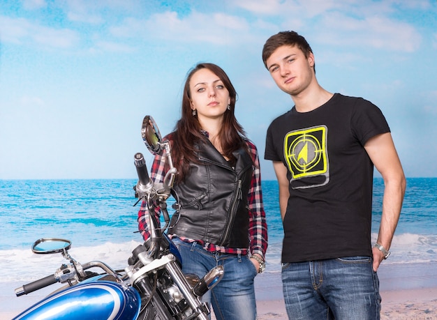 Молодая белая пара в повседневных нарядах с мотоциклом, глядя в камеру, отдыхая на пляже