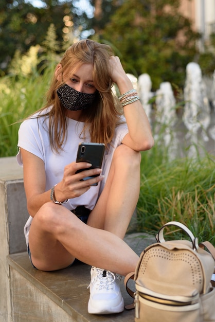 Молодая белая женщина в маске смотрит в свой телефон на улице