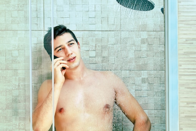 若い白人男性がシャワーで水の流れの下に立っている間電話で話している