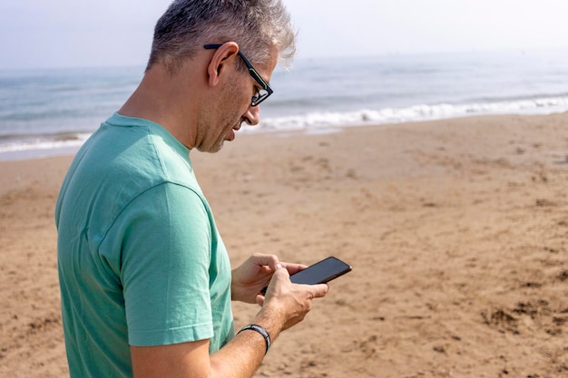 Young whitehaired man looking at his cell phone while walking on the beach