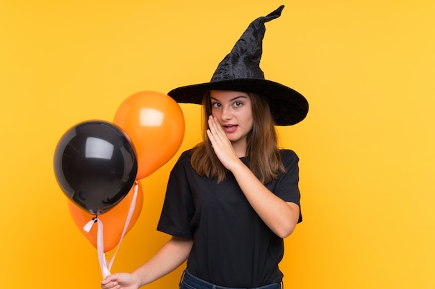 何かをささやくハロウィーンパーティーのための黒とオレンジの気球を保持している若い魔女