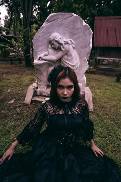 A young witch in a long black dress and scary makeup was sitting in front of the tombstone