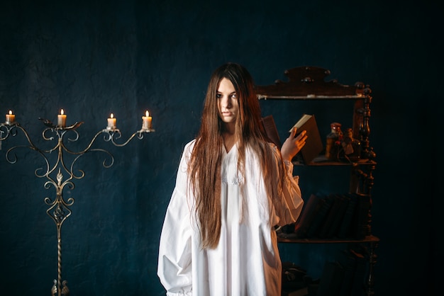 Young witch in white shirt holds spellbook in hands, candles. Dark magic ritual, occultism and exorcism, witchcraft