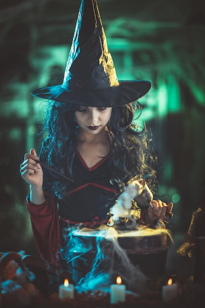 화난 사악한 얼굴을 한 젊은 마녀는 한 손에는 마법의 물약이 든 잔을, 다른 한 손에는 마술 지팡이를 들고 있습니다.